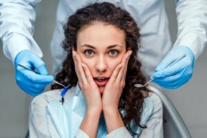 Patient in shock in dentist’s chair with hands pressed to her face.